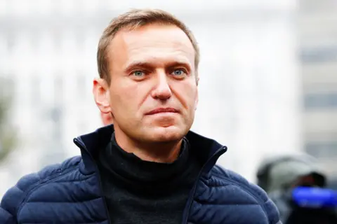 Getty Images Alexei Navalny has light hair and clear blue eyes in this photo and appears to be staring boldly at something beyond the camera. The photo is taken outside on what appears to be a cold morning. He has a dark sweater on under a blue puffa jacket, unzipped.