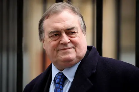 Getty Images John Prescott is pictured looking slightly over his shoulder with a wry expression on his face. He has dark hair, slightly greying, and is wearing a navy blue overcoat over a suit. His shirt is light blue and his tie is dark blue.