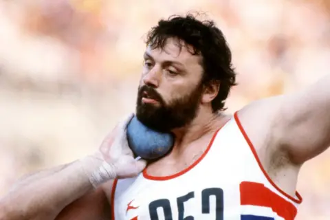 Alamy A picture of Geoff Capes competing in an outdoor event, holding a shot under his chin as he prepares to put it. He has a thick beard and is wearing a white-and-red athletics vest.