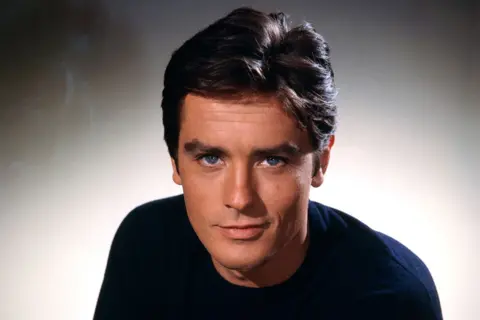 Getty Images Undated 1960s publicity photo of Alain Delon (in colour) when he would have been in his late 20s or early 30s. He has dark, slightly bouffant hair,  clear blue eyes and is smiling slightly. He wears a black sweater and is looking straight at the camera. 