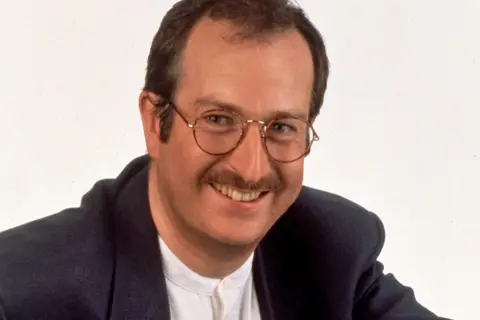 Publicity shot of Steve Wright against a white background. He has thinning dark hair, glasses and a moustache and is smiling broadly for the camera. He wears a white buttoned-up collarless shirt and a dark jacket.