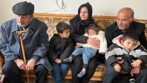 Aamir Peerzada/BBC A woman, three young children and two older men are seen sitting on a bench.