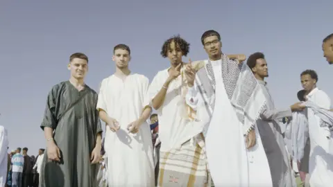 Kids of the Colony A picture of Zak Hajjaj, Anas Omar, Abu Finiin, and Kayum Miah standing in a crown of people dressed in a mix of Muslim and Somali cultural attire.