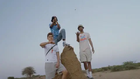 Kids of the Colony Zak Hajjaj, Abu Finiin, Kayum Miah standing next to a large ant hill with Abu standing on top