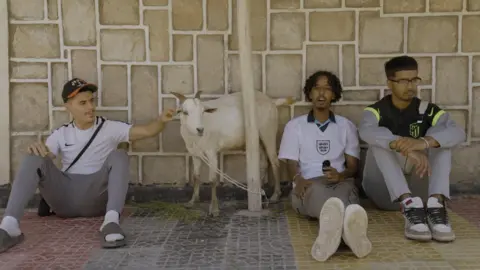 Kids of the Colony Zak Hajjaj, Abu Finiin, Kayum Miah sitting down against a wall with Zak petting a goat between himself and Abu