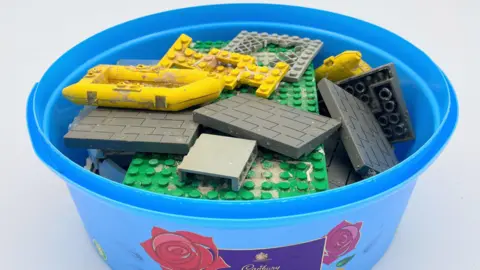 Tracey Williams A blue plastic sweet tin full of found Lego pieces including two yellow life rafts, a large green model base and grey roofing tiles.