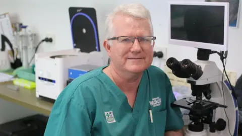 Tiffanie Turnbull/BBC A man with glasses and white hair in green scrubs looks at the camera and smiles. He is sitting in front of a desk with medical equipment including a microscope on it.