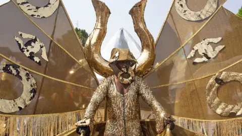 Emmanuel Adegboye / EPA A man in a gold sequin outfit with a giant manilla behind him holding up gold wings takes part in a carnival in Calabar, Nigeria