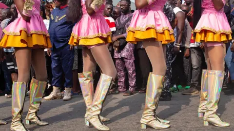 Emmanuel Adegboye / EPA Parade onlookers look through the legs of performers in gold boots and pink and orange