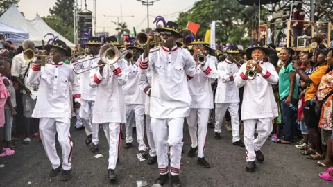 Olympia De Maismont / AFP A group of men dressed in white outfits and black large brimmed hats blow trumpets as they parade down a street in Calabar, Nigeria