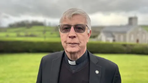 Fr Sean Moore wearing dark glasses, dark jacket, black shirt with white clerical collar. He is standing in a field - in the distance behind him the stone building of a church can be seen.