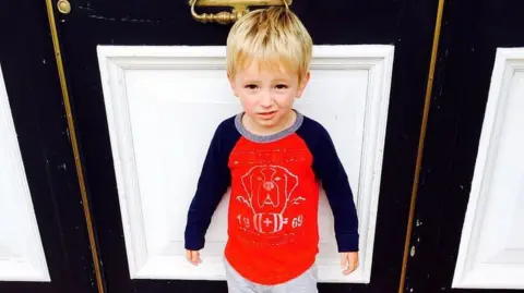 James Hunt Tommy in a red top as a young boy, standing in front of a door.