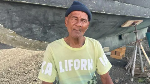 Charles Carter poses in front of his boat. He is wearing a blue knitted cap and a pale yellow T-shirt with the letters California on it in white.