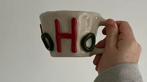 Mollie Warrington A hand, with a grey sleeve, holds up a mug, beige in colour with a bright red upper case 'H' and a lower case, green 'o'.