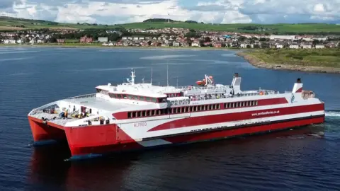 Christopher Brindle A red and white large catamaran with the name Alfred on the side, pictured in the foreground with a small seaside town in the background. 