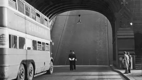 Getty Images Archive black-and-white photo from 1946, showing a double-decker bus paused on Tower Bridge. The section of the road which raises to let boats through is up and looks like a steeply sloping wall in front of the bus. Two policemen chat in the road, wearing helmets and arm gaiters. Two men in suits, one carrying a coat, are on the pavement and are looking toward the section of raised bridge