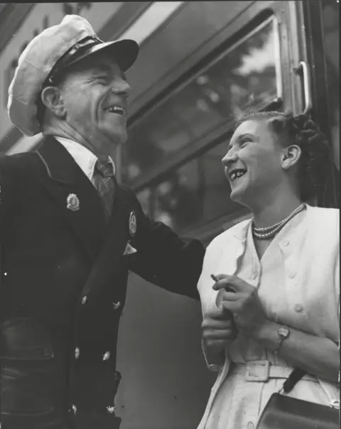 ANL/Shutterstock Albert Gunter and May Walshaw facing each other laughing and standing by a bus. Mr Gunter is wearing his bus uniform and is on the left of the picture, looking right and down. Miss Walshaw is on the right and is looking left and up. Miss Walshaw is wearing a pale dress with a matching short jacket and a double string of pearls. She has a handbag over her left elbow and holds a cigarette in her left hand.