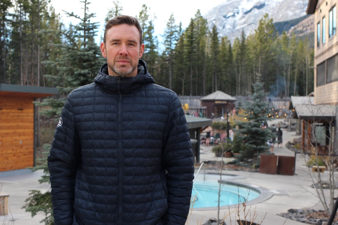 A man in a black puffer jacket poses for a photo at an outdoor Nordic spa in Kananaskis, Alta. with mountains in the background.