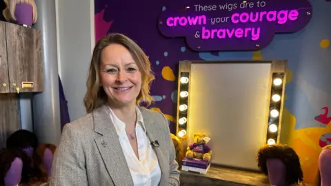 A woman with dark blonde hair down to her neck stands smiling in front of a theatre-style make-up mirror with lights down each side. She is wearing a white blouse and a grey jacket. A sign above the mirror reads: "These wigs are to crown your courage and bravery."