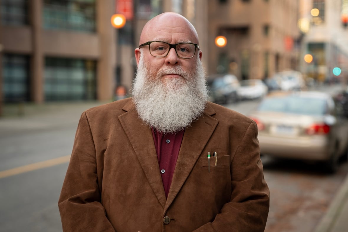 A bald man stands in the road in a brown jacket with a full, white beard and glasses.