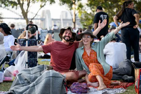 Bianca De Marchi/AAP Image via REUTERS Revellers gather at Mrs. Macquarie's Point ahead of the New Year's Eve celebrations in Sydney, Australia, 31 December 2024. A man in a brown t-shirt and a woman in an orange dress and grey shirt, both wearing hats, sit on the ground in a crown and wave to the camera
