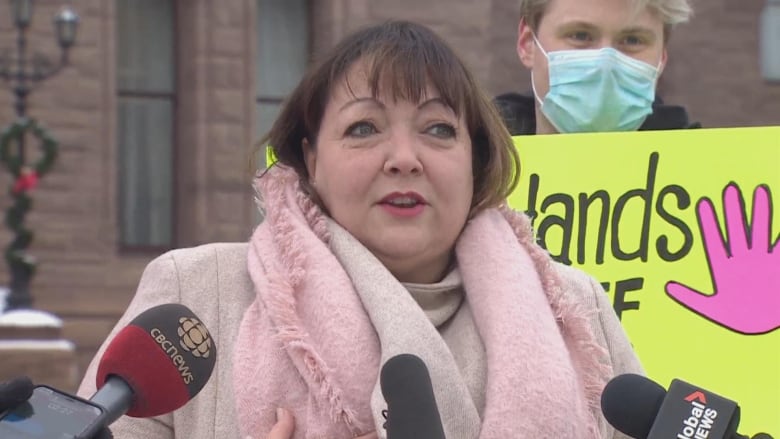 Woman in pink coat and scarf stands in front of brown building, speaking into microphones