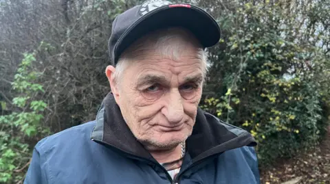 BBC A man with white hair and stubble stands in front of woodland. He is wearing a black baseball cap and blue raincoat. 