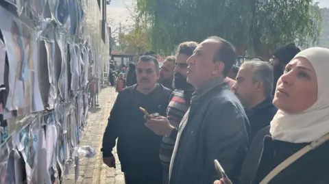 BBC Family members and friends of missing prisoners stare at a wall of printed photographs of dead men posted on a wall outside Mustahed Hospital in Damascus.