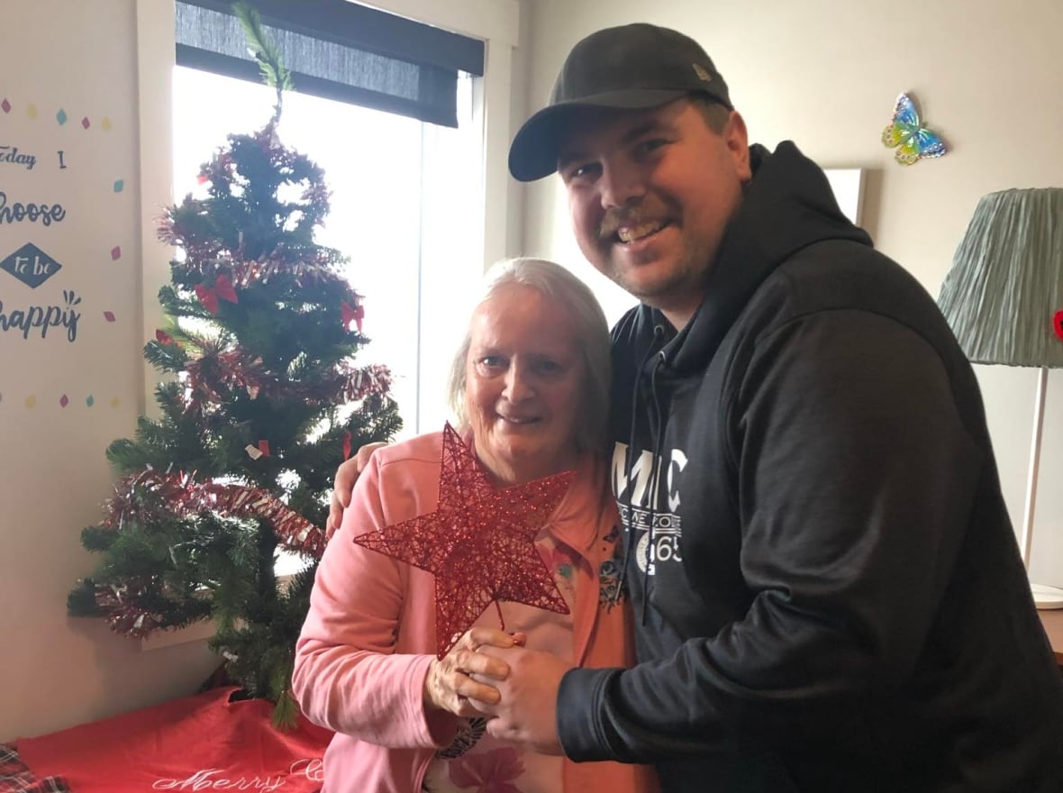 A white man wearing a black hoodie and ball cap, is hugging a shorter, older woman, wearing a pink sweater. Together, they are holding a red star Christmas tree topper in front of them.