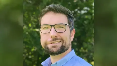 Family photo Aaron Jones with dark hair and glasses smiling at the camera. He is standing in front of a bushes