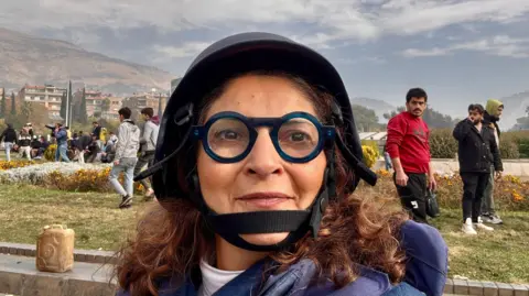 BBC Lina Sinjab, a Syrian BBC journalist with brown hair and with thick-rimmed dark blue circular glasses, wearing a press helmet, having returned to Damascus after the fall of the Assad regime in 2024