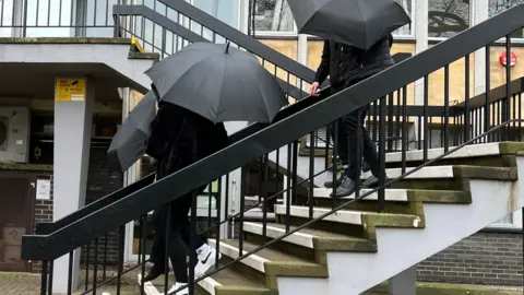 BBC Three people walking down a set of steps holding umbrellas. They are all wearing black clothing. 