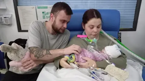 Irwin Mitchell Adyn Humphries sits with partner Chelsea Wootton as she holds their daughter Ava-Lea, who is attached to medical equipment, as they sit on a chair in a hospital ward.