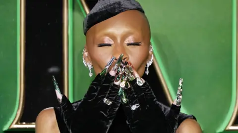 Tolga Akmen/EPA Cynthia Erivo on the green carpet at the London Premiere of Wicked. The picture is close up of Erivo's face. She has her eyes closed and is blowing a kiss to the camera. She is wearing black velvet gloves and you can see her long green fingernails.