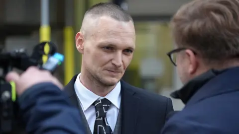 BBC James Howells listening to reporter who is facing him, with a camera man to the left of the reporter. Mr Howells is wearing a white shirt and black tie with a white pattern and a grey checked waistcoat and navy blazer. 