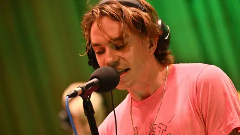 BBC Sports Team in session for BBC 6 Music. The photo shows vocalist Alex Rice at Maida Vale Studios, singing into a microphone while wearing headphones.