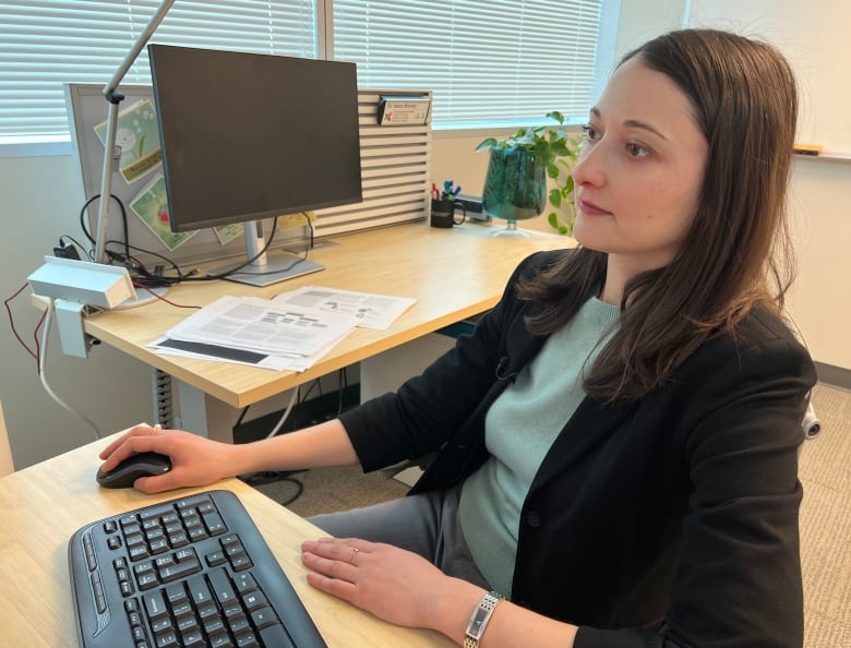 Profile of a woman with brown hair past her shoulders wearing a black blazer sits at computer using the mouse. 