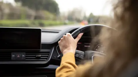 Getty Images A driver behind the wheel of a car with the dashboard in view.