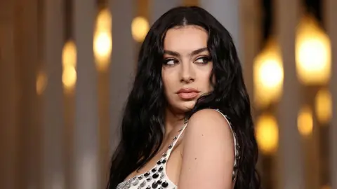 Getty Images Charli XCX wearing a white dress with shiny tear drop sequins on. She is facing the camera but looking to her left away from the lens. She has long black hair and make up on