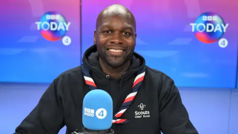 BBC Chief scout Dwayne Fields in the Today studio. A light blue Radio 4 microphone can be seen in the foreground in front of him and the Today logo can be seen on screens in the background. Fields is wearing his scout necker on top of his black hoodie which has the Scouts logo and his title printed on the left breast.