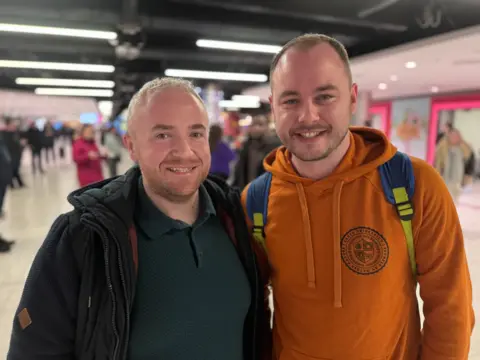 Craig Ballentine pictured in a camel coloured hooded top with his friend Sean who is wearing a dark top and jacket at the airport. They are smiling at the camera