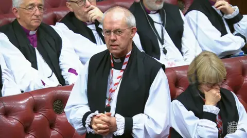 UK Parliament/PA Archbishop Justin Welby stands, hands clasped, in the House of Lords chamber wearing black and white robes.