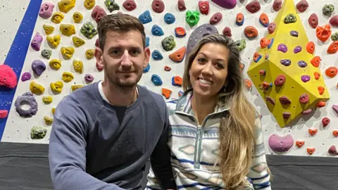 Kellie Burston A couple wearing athletic clothing sit on a mat in front of an indoor bouldering wall