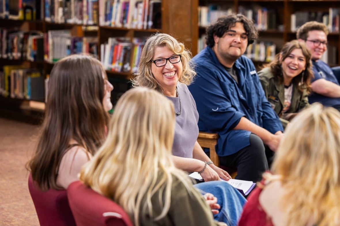 A woman with blonde hair and black framed glasses is smiling at a group of teens that are in discussion with her. 