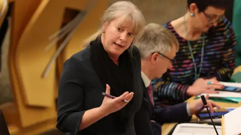 PA Media Shona Robison, with blonde hair and wearing a black dress, stands to speak in the Scottish Parliament with a pen in her hand 