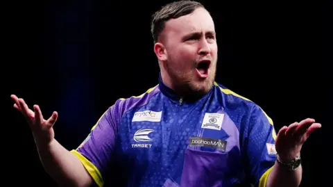 PA Media Darts player Luke Littler wearing his purple and yellow playing top holds out his hands and encourages the crowd