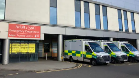 BBC Three ambulances parked outside the Queen Elizabeth University Hospital. A sign of the building reads Adults A&E, emergency department and major trauma centre.