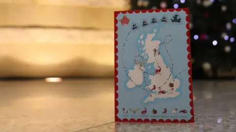 GCHQ A map of the United Kingdom with a Christmas-themed design, showing a trail of Santa’s sleigh and various holiday icons such as reindeer, robins, and other animals, held in front of a decorated Christmas tree with ornaments