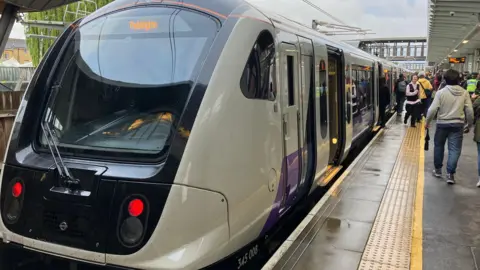 BBC An Elizabeth line train at the platform with its doors open. There are people walking along the platform beside the train. 