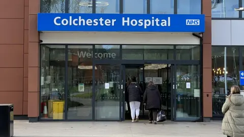 Simon Dedman/BBC The entrance to Colchester Hospital. The name of the hospital along with the blue NHS logo sit above the doors. Patients can be seen walking through the doors.